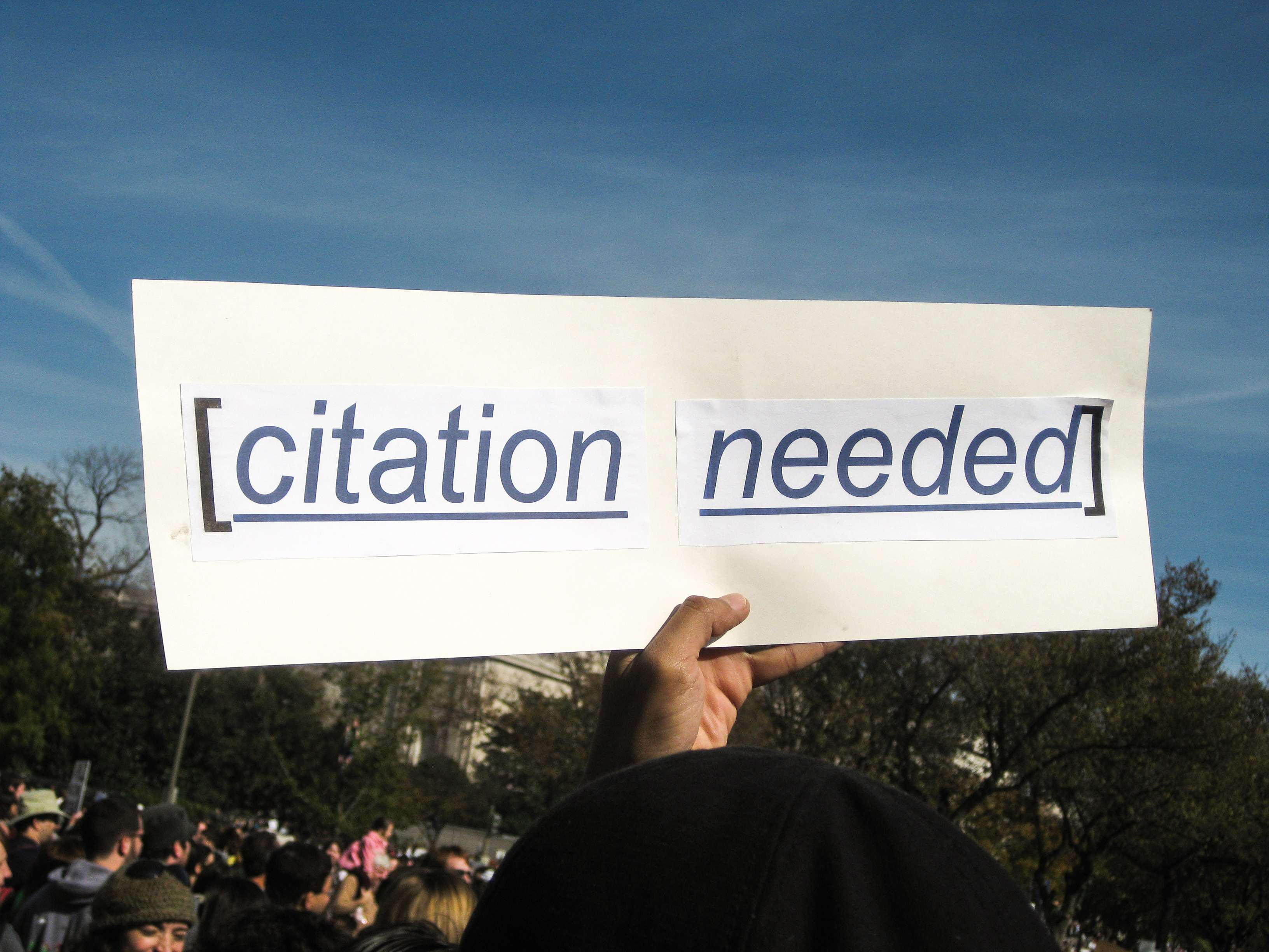 Image of a sign held up by a hand in the air that states in brackets citation needed. In the background is a large number of people with a blue sky.